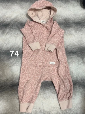Rosa blommig byxdress från Newbie - Mysig rosa byxdress från Newbie med blommigt mönster och huva. Muddar vid ärmar och benslut samt praktisk dragkedja längs sidan. Perfekt för chill dagar hemma eller när du vill vara extra bekväm.🌸 Fint använt skick. Storlek 74
