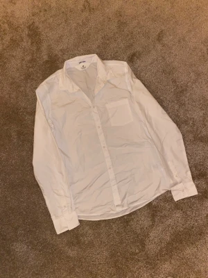Vit skjorta från H&M med knappar - Klassisk vit skjorta från H&M med lång ärm och knappar hela vägen framtill. Skjortan har en bröstficka och är tillverkad i ett lätt och bekvämt bomullsmaterial. Perfekt för en clean och stilren look.