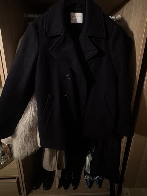 Svart skepparkavaj från Selected Femme - Klassisk svart skepparkavaj från Selected Femme med bred krage och dubbelknäppning. Jackan har två snedställda fickor framtill och är tillverkad i ullblandning. Perfekt för dig som vill ha en tidlös och stilren look.