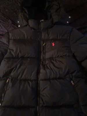 Svart pufferjacka från Polo Ralph Lauren - Säljer en svart pufferjacka från Polo Ralph Lauren med röd broderad logga på bröstet. Jackan har huva, dragkedja framtill och praktiska fickor med dragkedja. Perfekt för kalla dagar och riktigt snygg streetstil.