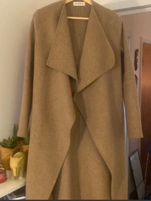 Beige omlottkappa från Wera - Säljer en snygg beige kappa från Wera med omlott-design och bred krage. Jackan är lång och har en stilren look som passar perfekt till lager-på-lager. Materialet är mjukt och känns lyxigt, och ärmarna är långa för extra värme.