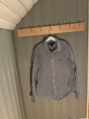 Randig skjorta från Ralph Lauren - Säljer en klassisk skjorta från Ralph Lauren med blåvita ränder och broderad logga på bröstet. Skjortan har lång ärm, knappar framtill och är tillverkad i mjuk bomull. Perfekt för dig som gillar stilrena och tidlösa plagg.