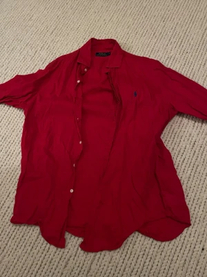 Röd linneskjorta från Polo Ralph Lauren - Säljer en röd skjorta från Polo Ralph Lauren i linne. Skjortan har klassisk krage, knappar framtill och den ikoniska blå loggan broderad på bröstet. Perfekt oversized fit för en chill look.