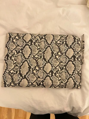 Datorfodral Gina Tricot - Snygg datorfodral från Gina Tricot i svart och beige ormmönster. Väskan har dragkedja upptill och är tillverkad i syntetmaterial. Perfekt storlek för det viktigaste och ger en cool touch till din outfit.