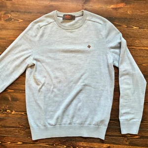 Ljusblå  merinoulltröja från Morris s.S - Säljer en stilren ljusgrå tröja från Morris i 100% merinoull. Tröjan har rund halsringning, diskret brunt broderat emblem på bröstet och ribbade muddar. Perfekt för dig som gillar klassisk och clean stil.