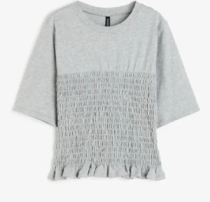 Ljusgrå topp med smock och volang - Supersöt ljusgrå topp från Divided med smockad front och volangkant nedtill. Toppen har rund halsringning och halvlånga ärmar. Tillverkad i mjuk bomull som känns skön mot huden. Perfekt till jeans eller kjol för en chill look. Har ett märke på baksida arm.