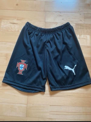 Portugal Puma svarta träningsshorts - Säljer ett par svarta träningsshorts från Puma med Portugals landslagsemblem på ena benet och Puma-logga på det andra. Shortsen har elastisk midja och är tillverkade i ett lätt och svalt syntetmaterial, perfekta för fotboll eller träning.