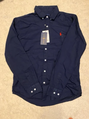 Mörkblå skjorta från Ralph Lauren - Klassisk mörkblå skjorta från Ralph Lauren med vit knappstängning och ikonisk röd logga broderad på bröstet. Skjortan har lång ärm, krage och är tillverkad i mjuk bomull. Perfekt för dig som vill ha en stilren och tidlös look. Priset kan diskuteras😊