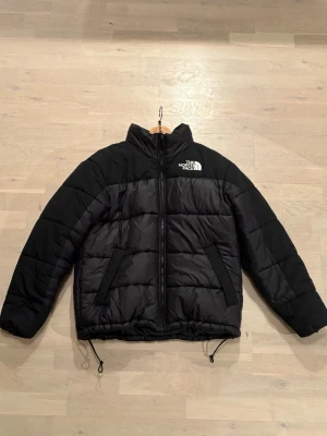 Svart pufferjacka från The North Face - Säljer en svart pufferjacka från The North Face i storlek M. Jackan har hög krage, två sidofickor med dragkedja och justerbar dragsko nedtill. Klassisk logga på bröstet och ryggen. Perfekt för kalla dagar och riktigt snygg till streetwear.