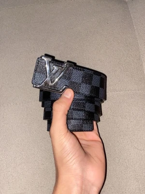 Rutig svart bälte - Säljer ett bälte i svart med klassiskt rutigt mönster och stor logga i silver på spännet. Bältet är gjort i skinn och har en snygg kontrast med ljus insida. Perfekt accessoar för att lyfta din outfit.