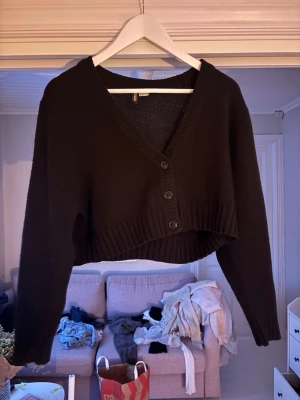 Svart croppad kofta med knappar - Svart croppad kofta från H&M med v-ringning och tre knappar framtill. Tröjan har långa ärmar och bred ribbstickad kant vid ärmslut och nederkant. Perfekt att slänga på över ett linne eller t-shirt.