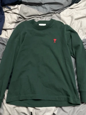 Mörkgrön långärmad tröja från Ami - Snygg mörkgrön långärmad tröja från Ami Alexandre Mattiussi med röd broderad logga på bröstet. Tröjan har rund halsringning och är gjord i mjuk bomull. Perfekt för dig som gillar stilrena plagg med en twist.