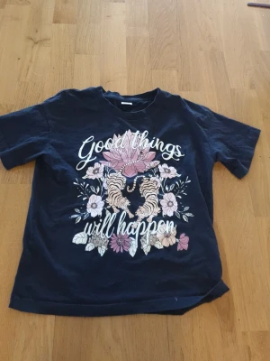Blå t-shirt med tigrar och blommor Lindex - Mörkblå t-shirt från Lindex med tryck av två tigrar, rosa och vita blommor samt texten 'Good things will happen' på framsidan. Klassisk passform och rund hals. Tillverkad i mjuk bomull, perfekt för en chill look.