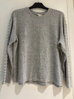 Grå tröja med pärldetaljer - Mysig grå tröja med rund hals och långa ärmar. Ärmarna är dekorerade med pärlor längs hela längden för en extra snygg touch. Perfekt för dig som vill ha något enkelt men ändå med lite edge.