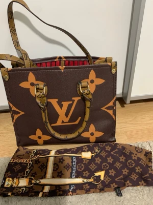 Handväska L.V - Säljer en snygg handväska L.V med klassiskt monogrammönster i brunt. Väskan har rejäla handtag med logodetaljer och röd- och svart randigt foder. Perfekt storlek för att få plats med allt du behöver och en scarf med kedjeprint medföljer. Väskan är använt sparsamt. 