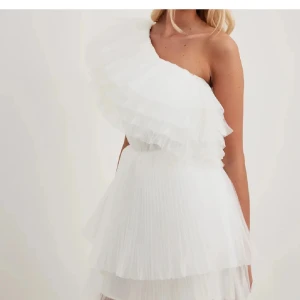Vit one shoulder klänning med volanger - Superfin vit klänning med one shoulder-design och stora volanger upptill. Klänningen har kort längd och flera lager av plisserat tyg som ger en fluffig och festlig känsla. Perfekt för student eller sommarens alla tillfällen! OBS klänningen är helt vit som på bilden men min egna bild är tagen i ett varmt ljus💕
