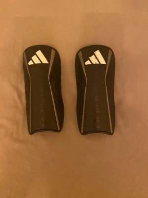 Adidas benskydd svart/vit - Säljer ett par svarta Adidas benskydd med vita detaljer och tillhörande svarta strumpor med tre vita ränder. Benskydden har en sportig design och är tillverkade i syntetmaterial, perfekta för fotboll. Strumporna är elastiska och sitter bra runt benskydden. Bara att skriva vid frågor och priset går såklart att diskutera!