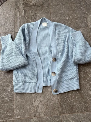Ljusblå stickad kofta från H&M - Supersöt ljusblå stickad kofta från H&M i storlek XS. Koftan har stora knappar framtill, ribbade muddar och en relaxed passform. Perfekt att slänga över en topp för extra mysfaktor. Klassisk modell med lång ärm och mjukt material.