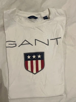 Vit GANT t-shirt med tryck - Vit t-shirt från GANT med stort tryck framtill. På bröstet finns GANT-loggan. Klassisk rund hals och korta ärmar. Knappt använd och från Kidsbrandstore! 
