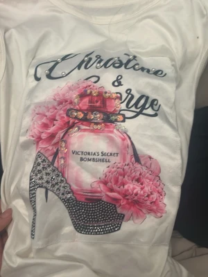 Vit t-shirt med blommor och paljetter - Vit t-shirt med tryck av parfymflaska, rosa blommor och en glittrig klack. Paljettdetaljer och pärlor på trycket ger extra bling. Texten 'Victoria's Secret Bombshell' syns på flaskan. Perfekt för dig som gillar statement-plagg.