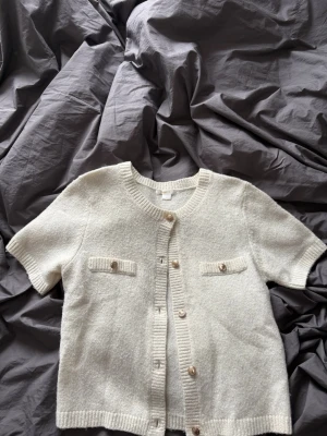 Kortärmad vit kofta med knappar - Säljer en söt kortärmad kofta i vit med ribbade kanter och dekorativa fickor framtill. Koftan har rund halsringning och stängs med flera knappar i matchande ton. Perfekt att slänga över en topp för en chill look. Säljer den då den inte kommer till användning❣️