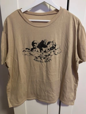 Beige t-shirt med tryck 'Angel Baby' - Beige t-shirt i bomull med svart tryck av änglar och texten 'ANGEL BABY' på bröstet. Modellen är loose fit med korta ärmar och rund halsringning. Står inte storlek men skulle säga L