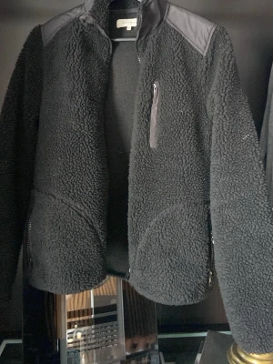 Svart teddyjacka från Studio Total - Mysig svart teddyjacka från Studio Total med dragkedjor på fickorna och en bröstficka med zip. Jackan har förstärkta axelpartier i syntetmaterial och en hög krage. Perfekt för dig som gillar stilren och chill look.