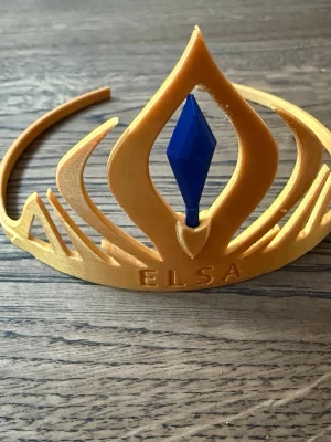 Elsa tiara i guld och blått - Säljer en tiara inspirerad av Elsa från Frost. Tiaran är i guldig plast med en blå diamantformad detalj i mitten och texten 'ELSA' framtill. Perfekt för cosplay eller temafest. Snygg och lätt att bära.