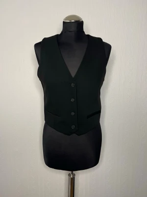 Svart kostymväst från H&M Oanvänd  - Stilren svart kostymväst från H&M i klassisk modell med fyra knappar framtill och två fickor. Västen har en djup v-ringning och är tillverkad i ett slätt tyg som ger en dressad look. Perfekt att styla över en skjorta eller t-shirt.