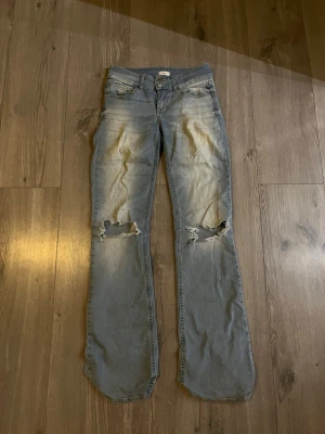 Ljusblå bootcut jeans med slitningar - Säljer ett par ljusblå bootcut jeans med slitna knän och snyggt tvättad look. Jeansen har klassiska fickor fram och två bakfickor med lock och knapp. Dock har de varit ett litet hål på höger ben på insidan lår som jag sytt ihop, inget som syns när man har dom på sig. Priset är inte hugget i sten!