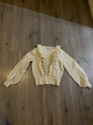 Vit stickad tröja med volang - Supersöt vit stickad tröja med volangdetaljer längs halsringningen och framtill. Tröjan har långärmade puffärmar och bred ribbad mudd i midjan. Perfekt för att lyfta din outfit. 