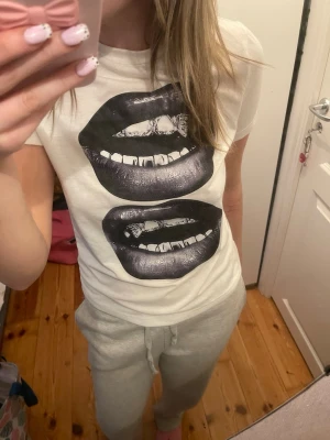 Vit t-shirt med läppar print - Säljer en vit t-shirt med coolt svartvitt tryck av två stora läppar på framsidan. T-shirten är kortärmad och har en klassisk passform. Perfekt för dig som vill sticka ut med en unik och edgy stil.