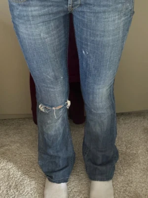 Levis 501 blå bootcut jeans - Säljer ett par klassiska Levis 501 jeans i blå tvätt med slitningar och hål på ena knät. Byxorna har låg midja, knappgylf och bootcut passform. Tydliga Levis-detaljer med synliga sömmar och logga på fickan. Perfekta för en avslappnad look.