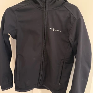 Svart vindjacka från Sail Racing - Svart vindjacka från Sail Racing med huva och dragkedja framtill. Jackan har stilren design med logga på bröstet och på huvan. Tillverkad i polyester, perfekt för blåsiga dagar. Snygg passform och praktiska fickor på sidorna.