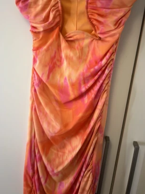 Orange och rosa långklänning Gina Tricot - Säljer en färgstark långklänning från Gina Tricot i storlek XS. Klänningen har ett coolt mönster i orange och rosa toner, med draperade detaljer längs sidorna och korta puffärmar. Materialet känns mjukt och stretchigt, perfekt för dig som gillar att sticka ut.