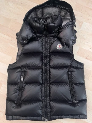 Svart dunväst från Moncler - Säljer en svart dunväst från Moncler med glansig finish och hög krage. Västen har två fickor med dragkedja, Moncler-logga på bröstet och quiltad design. Perfekt för lager-på-lager och har en stilren look som funkar till många outfits.