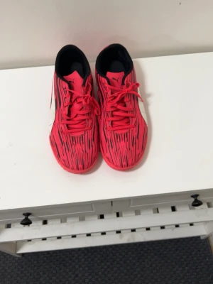 Röda fotbollsskor från Puma - Snygga fotbollsskor från Puma i röd färg med svarta och vita detaljer. Skorna har ett randigt mönster och klassisk snörning. Perfekta för dig som vill sticka ut på planen.