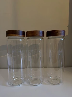 Tre glasburkar med acacia trälock - Säljer tre stilrena glasburkar med runda lock i mörkt trä. Perfekta för förvaring av pasta, ris, kaffe eller snacks. Burkarna är cylinderformade och transparenta så du ser enkelt innehållet. Locken har en snygg trälook som ger en modern känsla till köket.