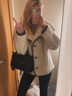 Vit teddyjacka med svarta detaljer - Mysig vit teddyjacka med svarta knappar och kontrasterande svarta kanter. perfekt för att hålla värmen under höst och vinter. Enkel att styla till både jeans och kjol.