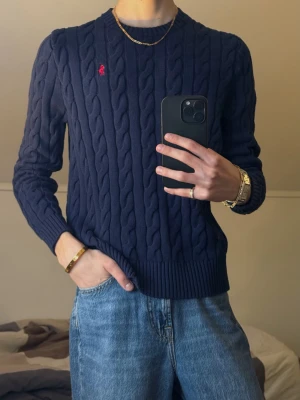 Mörkblå kabelstickad tröja Polo Ralph Lauren - Kabelstickad mörkblå tröja från Polo Ralph Lauren med rund halsringning och ribbade muddar. Tröjan har den klassiska röda logotypen broderad på bröstet. Perfekt för dig som gillar tidlös och stilren design.