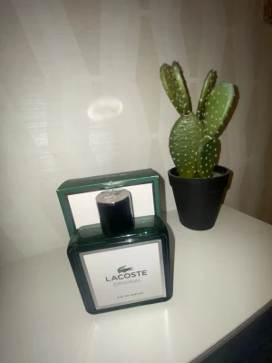 Lacoste Original Eau de Parfum 100ml - Fräsch parfym från Lacoste i en stilren, mörkgrön glasflaska med svart lock. Flaskan har en fyrkantig form och en enkel vit etikett med Lacoste-loggan. Perfekt för dig som gillar klassiska och sportiga dofter.