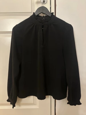 Svart blus med volangkrage - Säljer min jättefina svarta blus som är i nyskick. Den har lång ärm och volangdetaljer vid krage och ärmslut. Blusen har en lätt v-ringning framtill och kan både stylas öppen och stängd. Inga defekter eller liknande. 