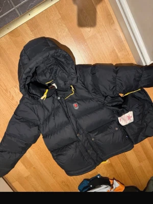 Fjällräven expedition lite  - Snygg svart dunjacka från Fjällräven med huva och gula detaljer på snörningen. Jackan har en quiltad design, stora fickor framtill och stängs med både dragkedja och knappar. Perfekt för kalla vinterdagar och riktigt varm.