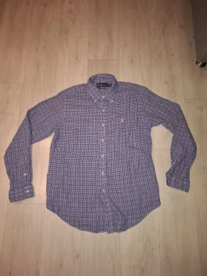 Stilren blårutig skjorta från Ralph Lauren i storlek L - Klassisk rutig skjorta från Ralph Lauren i blått och vitt med knappar framtill och button-down krage. Skjortan har lång ärm och en liten gul broderad logga på bröstet. Perfekt för en stilren och avslappnad look. Priset är självklart diskuterbart