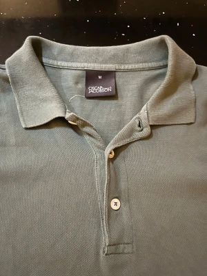 Grön pikétröja från Oscar Jacobson - Klassisk grön pikétröja från Oscar Jacobson med krage och två knappar framtill. Tillverkad i mjuk bomull som känns skön mot huden. Perfekt till jeans eller shorts för en avslappnad men stilren look.