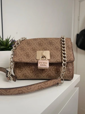 Brun handväska från Guess med kedja - Snygg brun handväska från Guess med logomönster och guldfärgad kedja. Väskan har ett elegant lås framtill med Guess-detalj och justerbar axelrem. Perfekt accessoar för att lyfta din outfit.