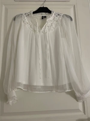 Vit blus med spets och volang - Vit blus från Vero Moda med spetsdetaljer vid halsringningen, volangkant och knytband framtill. Blusen har långa, transparenta ärmar och är tillverkad i ett lätt och luftigt polyestertyg. Perfekt för en chic och feminin look.