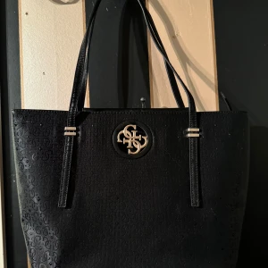 Svart handväska från Guess - Stilren svart handväska från Guess med präglat mönster och metallogo framtill. Väskan har dubbla axelband och rymlig insida, perfekt för att bära med sig allt man behöver. Snygga gulddetaljer vid handtagen ger en exklusiv känsla.