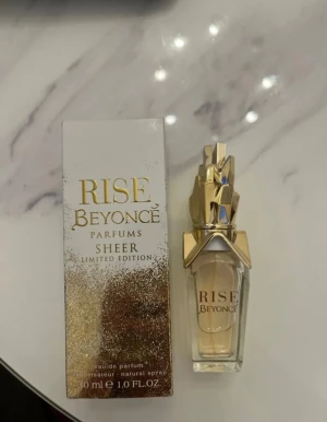 Beyoncé Rise Sheer Limited Edition - Rise Sheer Limited Edition från Beyoncé är en exklusiv parfym i en elegant glasflaska med guldiga detaljer och en unik, skulptural kork. Doften kommer i en 30 ml flaska och har en lyxig design med guld och vitt på förpackningen.