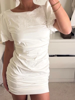Vit puffärmad miniklänning - Kort vit klänning med puffärmar och figurnära passform. Klänningen har en öppen rygg och är tillverkad i ett lätt, slätt material. Perfekt för sommarens alla tillfällen.